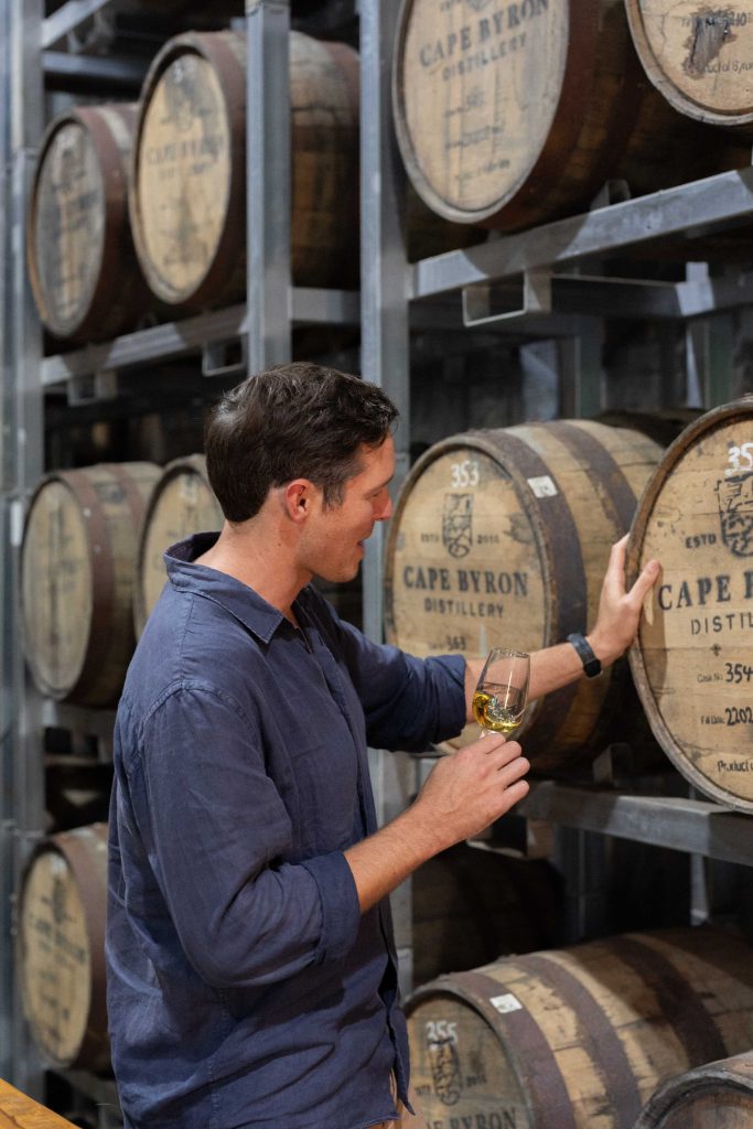man standing in front of wooden whisky barrels with a glass in right hand