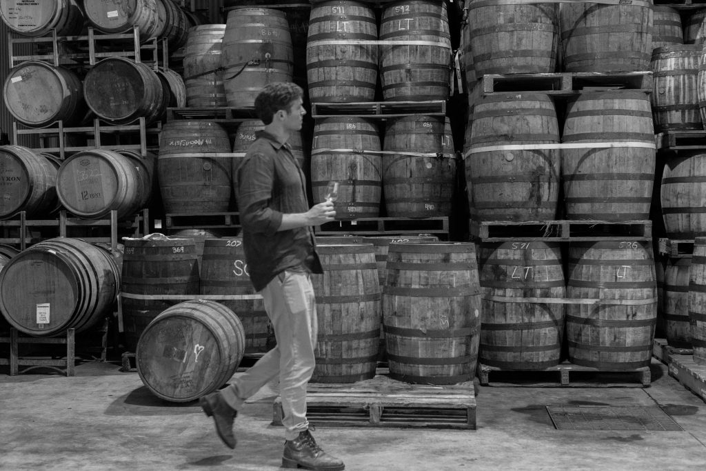 Black and white image of man walking in front of whisky barrels