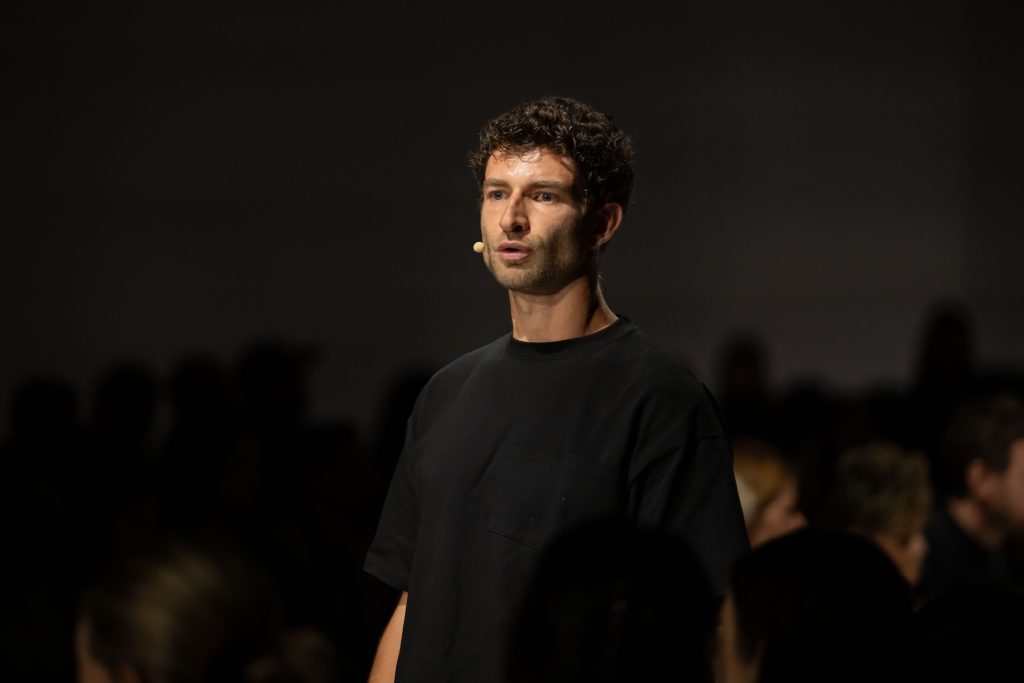 Rory Warnock standing with microphone on with black background room, talking to an audience.